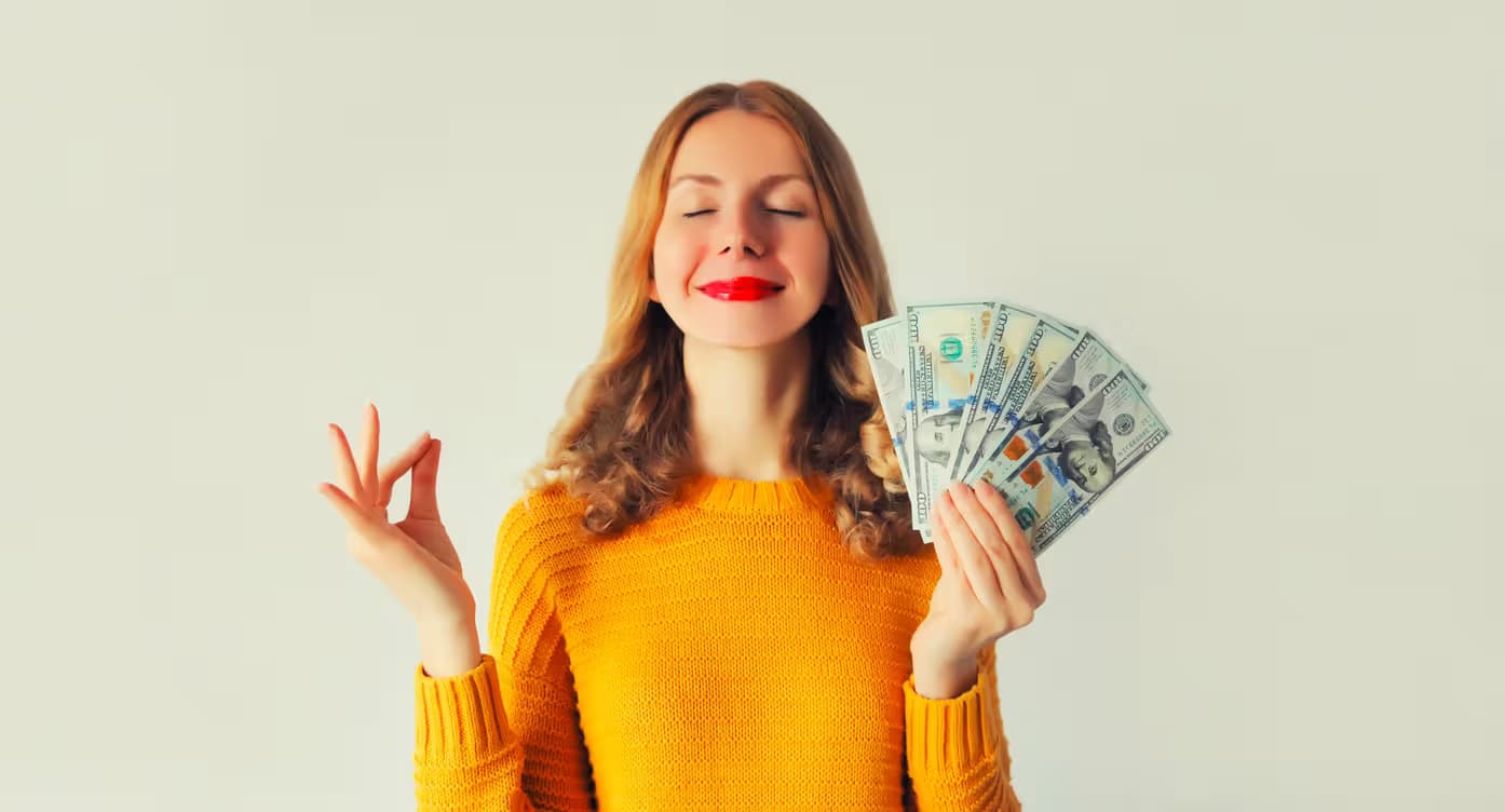 A smiling woman in a vibrant yellow sweater holding a fan of $100 bills, posing with a relaxed meditation gesture to represent the ease of earning passive income. The Lotto Nuts Referral Program