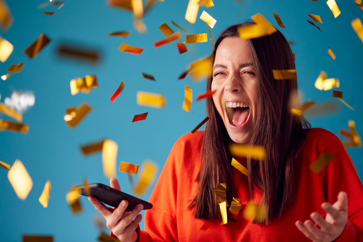 A joyful woman in a red sweater screaming with excitement while holding a smartphone as gold confetti falls around her, capturing the moment of a big lottery win or successful ticket purchase. How-To-Order-Lottery-Tickets-Online